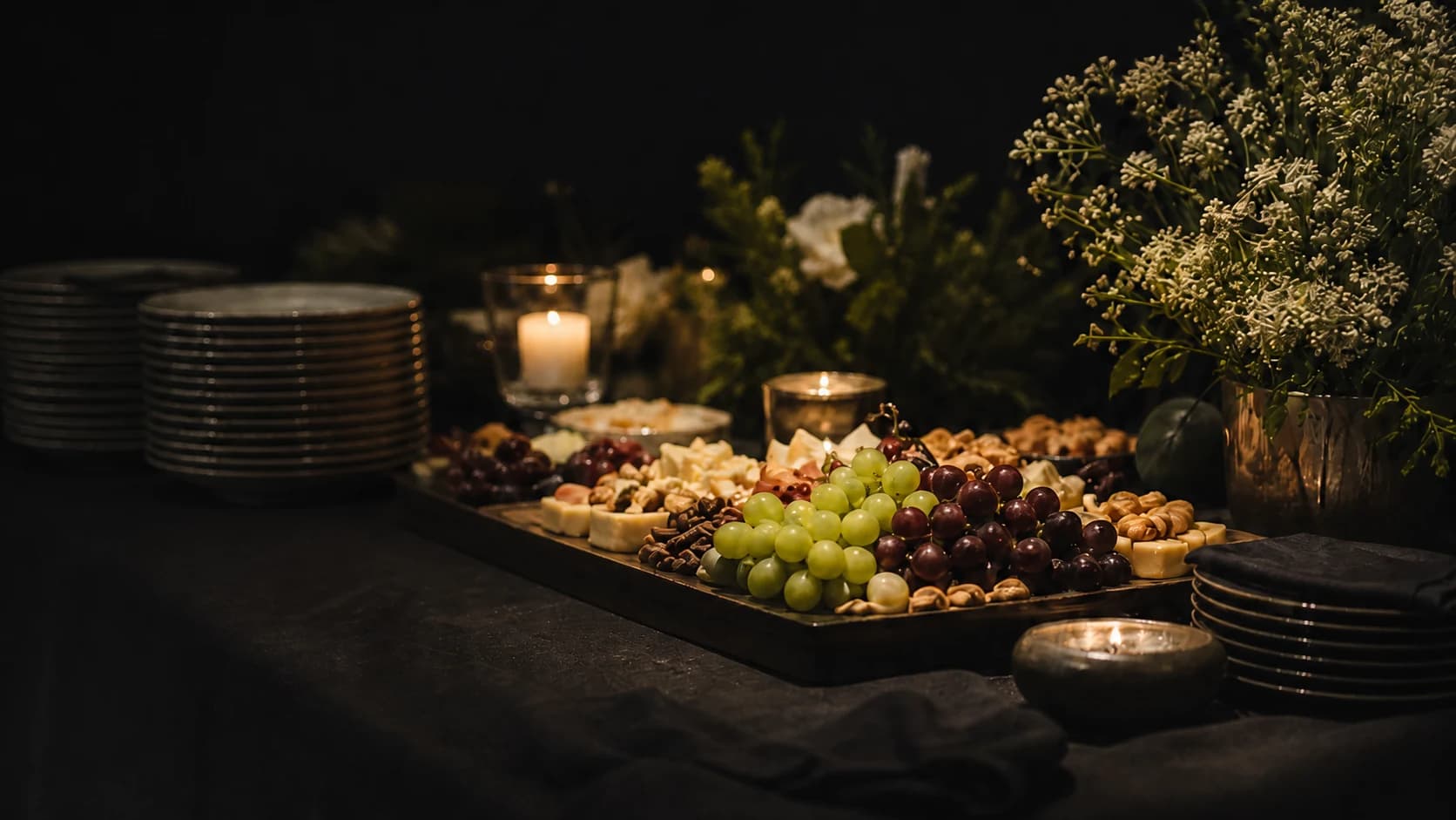 A candlelit catering table with grapes, cheeses, crackers, dark dinnerware, and cream-colored flowers