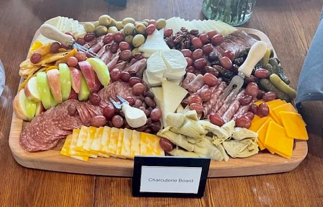 A labeled charcuterie board with cheeses, cured meats, grapes, olives, and crackers.