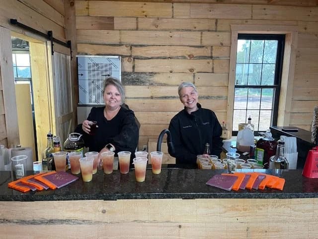 Two bartenders smiling behind a counter with prepared drinks.