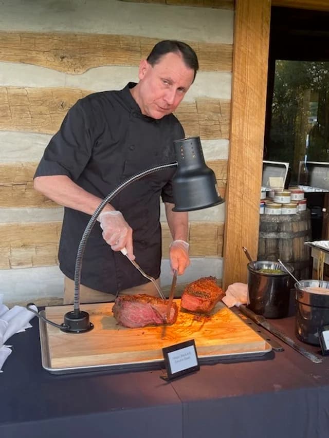 A chef carving a large cooked steak beneath a warming lamp.