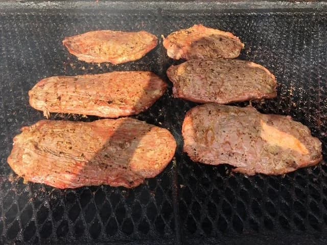 Six seasoned steaks cooking on an outdoor grill.